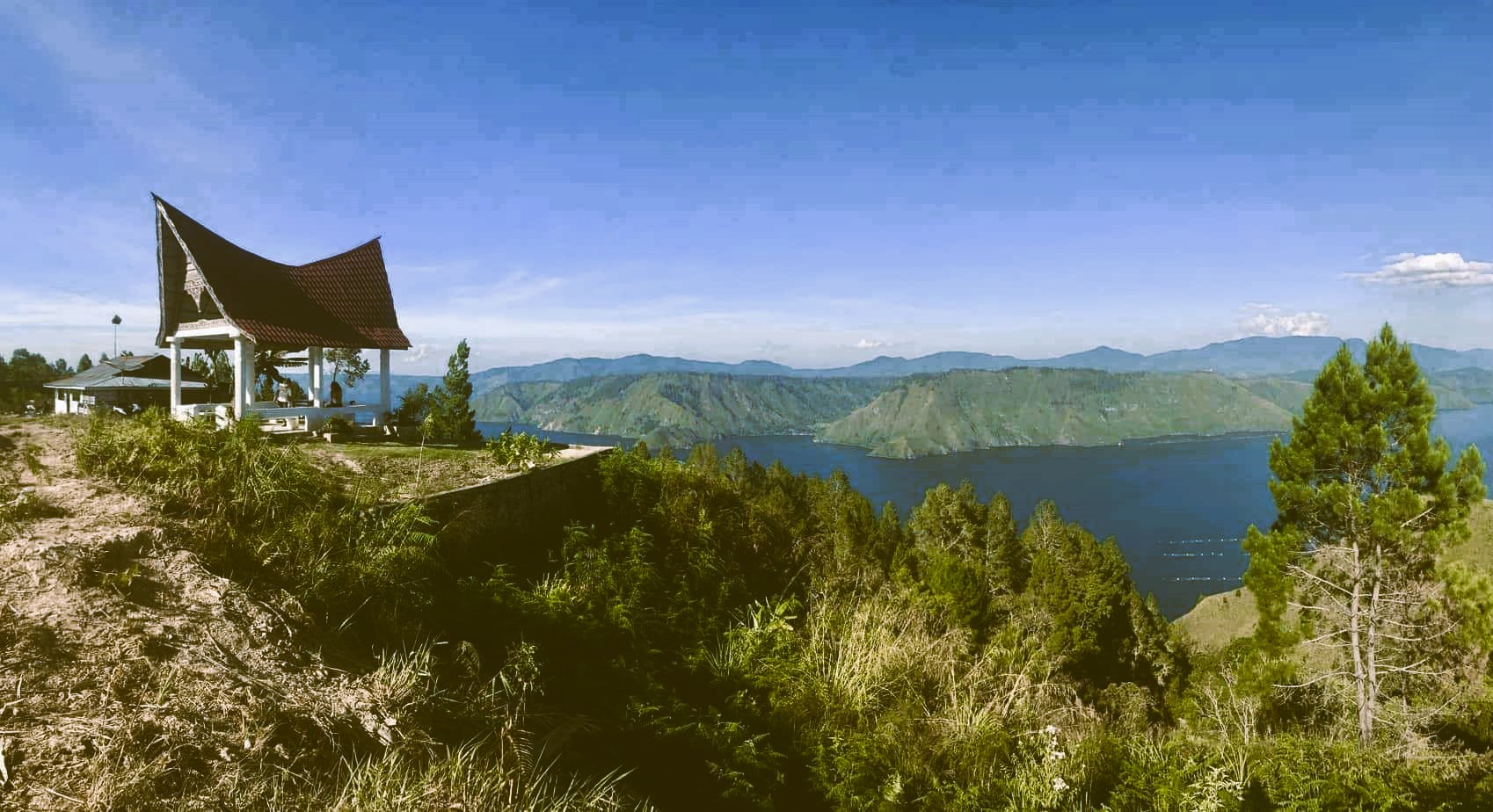 A view of lake Toba from a cliff, with a house and a Batak-style pavilion on the same cliff